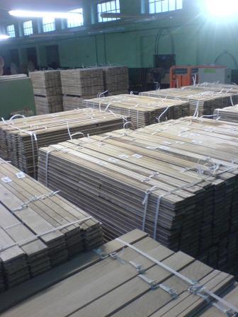 Stosy tarcicy budowlanej, deski podłogowe dębowe i sosnowe, spięte białymi taśmami, składowane w magazynie z oknami i zielonymi ścianami. Widoczne źródło światła w górnym, prawym rogu.